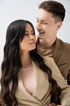happy man looking at stylish asian woman with long brunette hair isolated on grey