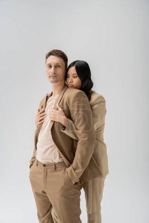 morena asiática mujer abrazando hombre de pie con las manos en bolsillos de pantalones beige aislado en gris