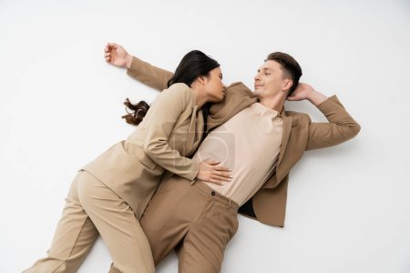 high angle view of fashionable interracial couple in suits lying and looking at each other on grey background