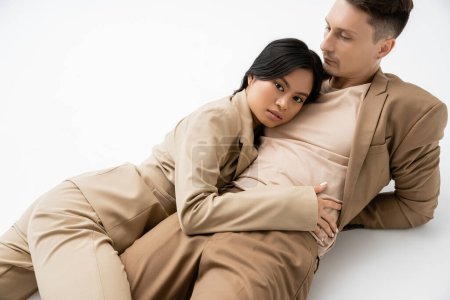 brunette asian woman in beige suit looking at camera while lying near trendy man on grey background