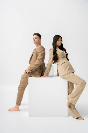 barefoot interracial couple in stylish casual attire posing back to back near white cube on grey background