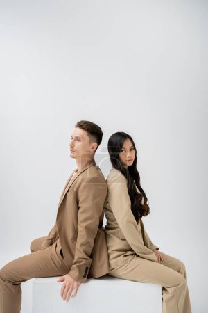 morena mujer asiática con el pelo largo mirando a la cámara mientras se sienta espalda con espalda con el hombre de moda aislado en gris