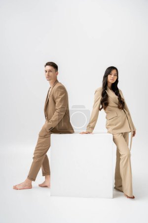 full length of barefoot multiethnic couple in beige pantsuits looking at camera near white cube on grey background