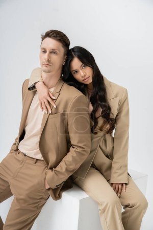 brunette asian woman hugging man in beige suit while sitting on cube and looking at camera on grey background