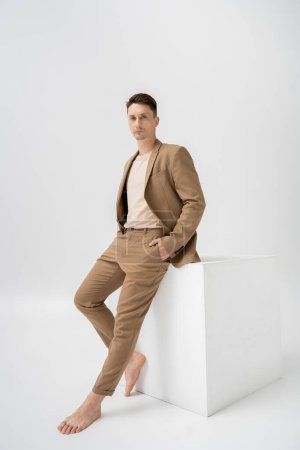 full length of barefoot man in beige pantsuit posing with hand in pocket near white cube on grey background