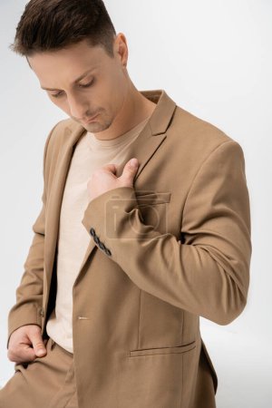 brunette man touching beige stylish blazer while posing on white background