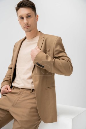 trendy man in beige suit looking at camera while sitting on white cube on grey background