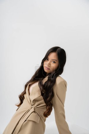 pretty asian woman with long brunette hair posing in stylish blazer and looking at camera isolated on grey