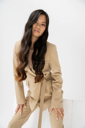young asian woman with long brunette hair wearing beige suit and looking at camera near cube on white background