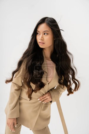 young asian woman with long brunette hair wearing beige jacket and looking away isolated on grey