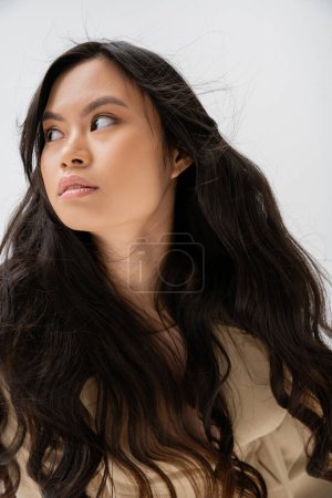 portrait of young asian woman with long brunette hair and natural makeup looking away isolated on grey