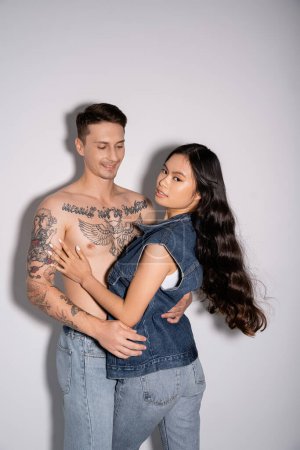 Téléchargez les photos : Musclé tatoué homme étreignant brunette asiatique femme regardant caméra sur gris fond - en image libre de droit