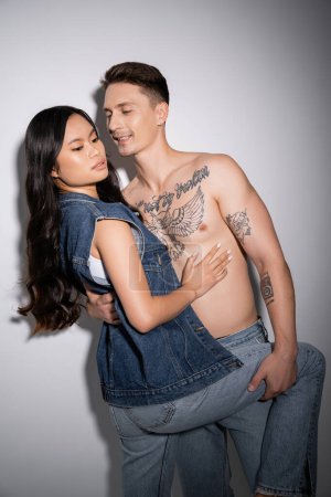 Photo for Smiling tattooed man embracing sensual asian woman in denim outfit on grey background - Royalty Free Image
