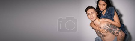 Photo for Tattooed and muscular man smiling at camera while piggybacking asian woman on grey background, banner - Royalty Free Image