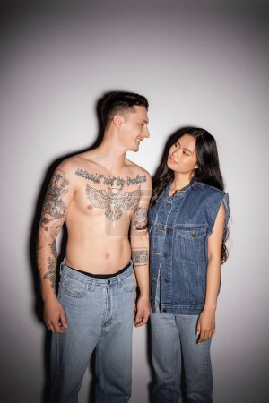 Téléchargez les photos : Torse nu tatoué homme et asiatique femme en denim vêtements sourire à l'autre sur fond gris - en image libre de droit