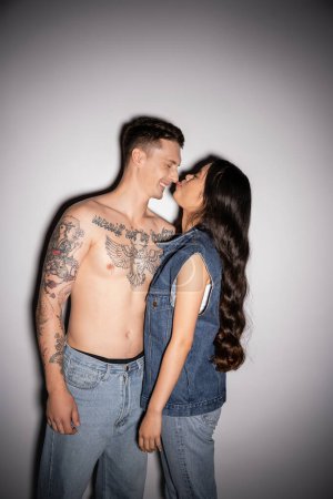 long haired asian woman sticking out tongue near tattooed man smiling with closed eyes on grey background
