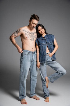 Téléchargez les photos : Full length of shirtless tattooed man and asian woman in jeans standing barefoot on grey background - en image libre de droit