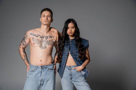 Téléchargez les photos : Stylish asian woman in denim clothes and shirtless man with tattooed body looking at camera on grey background - en image libre de droit