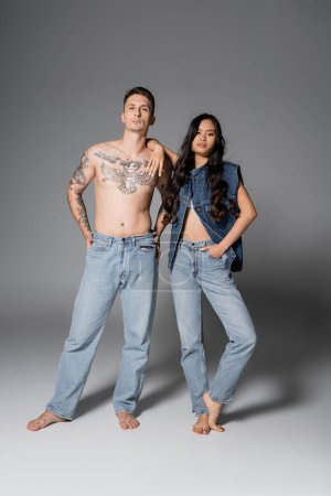 Téléchargez les photos : Full length of trendy asian woman and shirtless man with tattooed body posing in jeans on grey background - en image libre de droit
