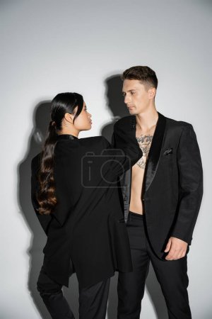 long haired asian woman and tattooed man in black elegant attire looking at each other on grey background