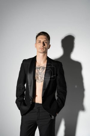 front view of shirtless tattooed man in black elegant pantsuit posing with hands in pockets on grey background with shadow