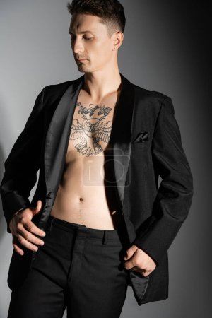 tattooed man wearing black elegant blazer over shirtless body posing on grey background