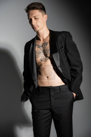 tattooed man in black stylish blazer on shirtless body posing with hands in pockets on grey background with shadow