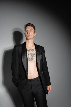 hombre tatuado sin camisa en blazer negro y pantalones mirando a la cámara sobre fondo gris con sombra