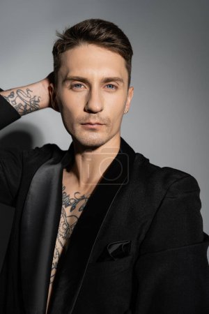 Foto de Stylish tattooed man in black blazer posing with hand behind head while looking at camera on grey background - Imagen libre de derechos