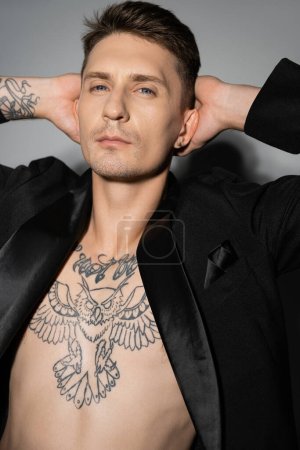 Foto de Hombre elegante con el cuerpo tatuado que lleva chaqueta negra y que sostiene las manos detrás de la cabeza sobre fondo gris - Imagen libre de derechos