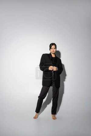 full length of barefoot asian woman wearing black stylish suit while looking at camera on grey background with shadow