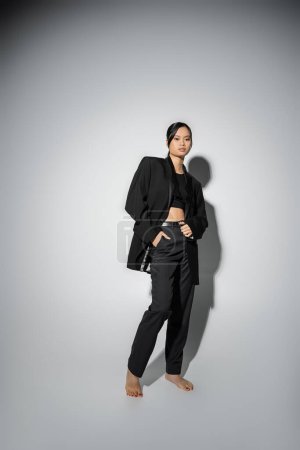 Foto de Full length of barefoot asian woman in black pantsuit posing with hand in pocket on grey background with shadow - Imagen libre de derechos