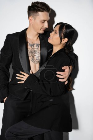 happy asian woman and tattooed man in black stylish attire looking at each other on grey background