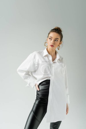 Foto de Trendy woman in white shirt and black pants posing with hand on hip isolated on grey - Imagen libre de derechos