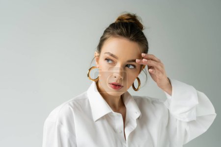 Foto de Portrait of young woman in white shirt and hoop earrings touching eyebrow and looking away isolated on grey - Imagen libre de derechos