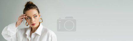 Photo for Portrait of pretty woman in hoop earrings and natural makeup looking away while touching eyebrow isolated on grey, banner - Royalty Free Image