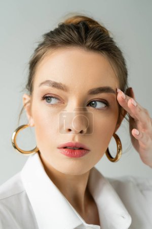 portrait of young woman with natural makeup and hoop earrings touching face and looking away isolated on grey