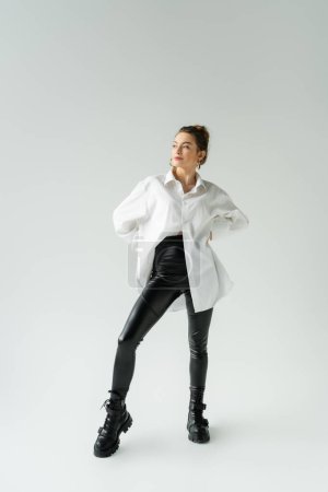 full length of woman in white oversize shirt and black tight pants posing with hands on waist on grey background