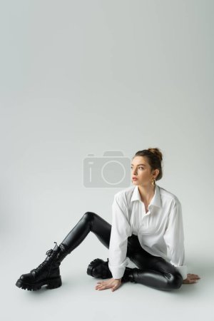 Foto de Full length of trendy woman in white shirt and black tight pants sitting and looking away on grey background - Imagen libre de derechos