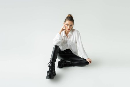 Foto de Full length of young model in black latex pants and rough boots sitting on grey background - Imagen libre de derechos