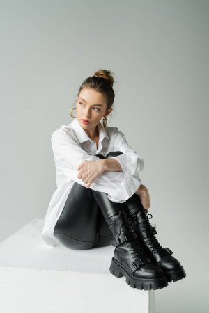 Photo for Full length of pretty woman in rough boots and white oversize shirt sitting on white cube and looking away isolated on grey - Royalty Free Image