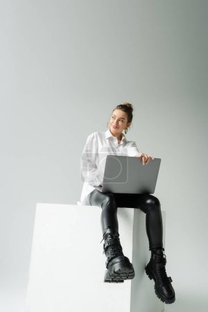 full length of happy woman in black latex pants and leather boots sitting on white cube with laptop on grey background