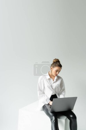 Photo for Young woman in oversize shirt and black tight pants working on laptop while sitting on cube on grey background - Royalty Free Image