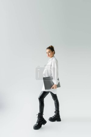 longitud completa de la mujer con estilo en pantalones de látex negro y botas ásperas posando con el ordenador portátil sobre fondo gris