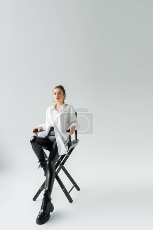 Foto de Full length of young woman in fashionable outfit sitting on chair and looking at camera on grey background - Imagen libre de derechos
