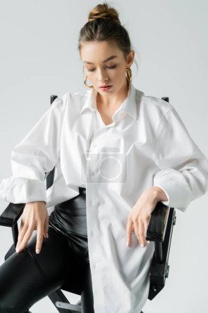 Foto de Stylish woman sitting on chair in white oversize shirt isolated on grey - Imagen libre de derechos