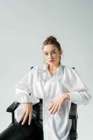 Foto de Stylish woman in white shirt and golden earrings sitting on chair and looking at camera isolated on grey - Imagen libre de derechos