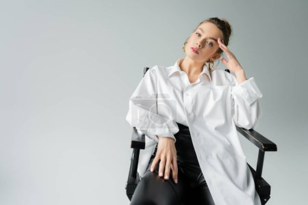 Foto de Pretty woman in white oversize shirt and black tight pants posing on chair with hand near head isolated on grey - Imagen libre de derechos