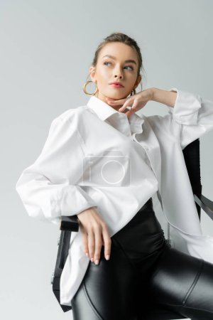 Foto de Young woman in oversize shirt touching chin and looking away while sitting on chair isolated on grey - Imagen libre de derechos