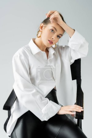 Foto de Stylish woman in hoop earrings and white oversize shirt posing on chair and looking at camera isolated on grey - Imagen libre de derechos